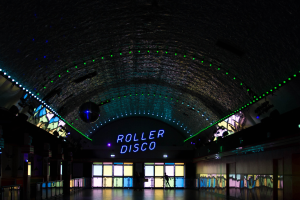 Ein Roller-Disco in einem Gebäude mit beleuchteter "Roller-Disco"-Anzeige, Menschen auf dem Boden stehend, Glastüren im Hintergrund und Säulen links und rechts.