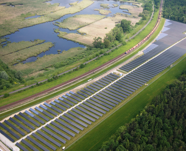 Luftaufnahme einer Solar-Farm mit Panelen, umgeben von Baumen, Gras und Wasser, mit einem Zug auf einer nahen Eisenbahnstrecke.