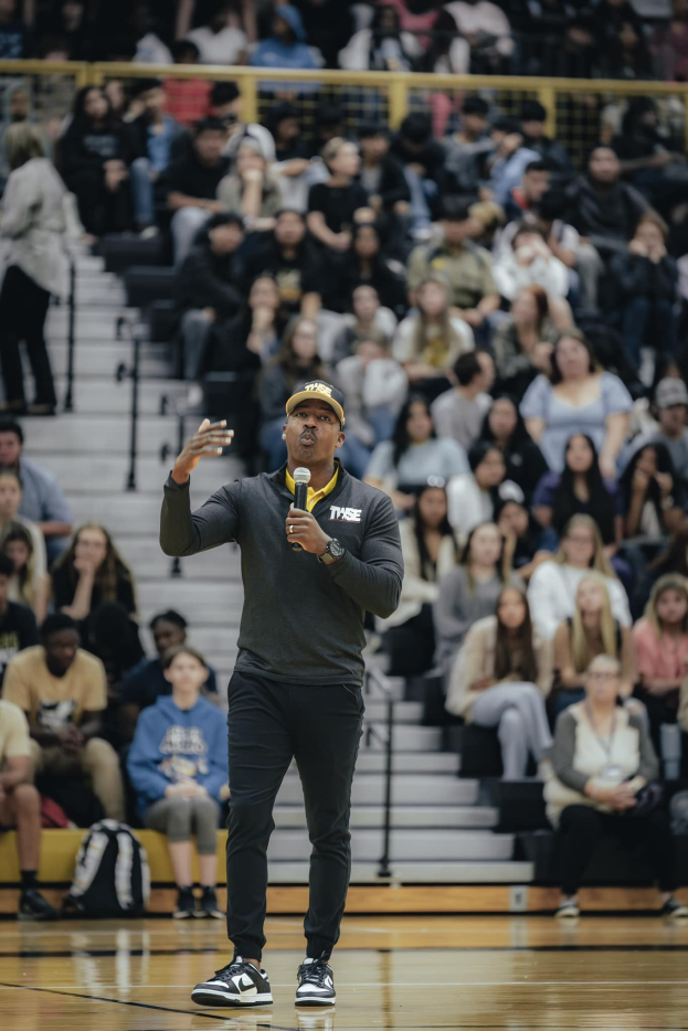 Ein Mann in einer Mütze und mit Mikrofon steht auf einem Basketballfeld, mit sitzenden Zuschauern und Taschen dahinter, wahrscheinlich der Cheftrainer während eines Spiels.