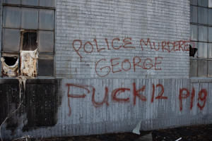 Ein Gebäude mit der Graffiti-Beschriftung "Polizei ermordet George Fuck 12 P19" an der Seite, das Fenster und verschiedene Gegenstände vorne zeigt.