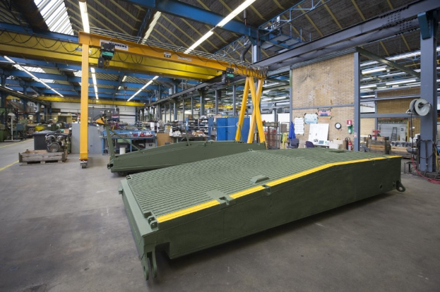 Ein große Lagerhalle mit zahlreichen grünen Behältern auf dem Boden, verschiedene Maschinen, Pfosten, Lichter und Stäbe, mit einer Wand im Hintergrund, die eine Uhr und angeklebte Papiere zeigt.