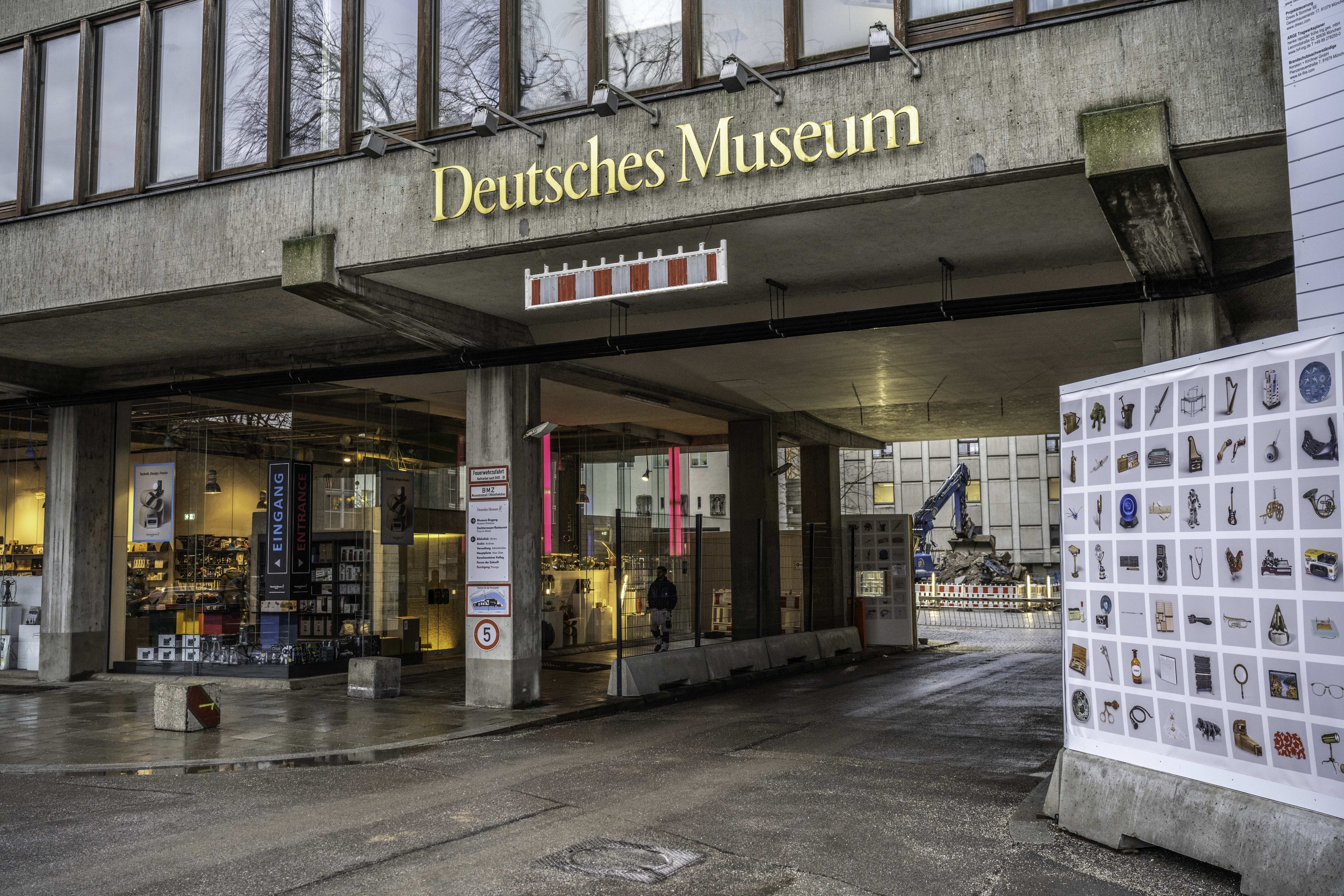 Außenansicht des Deutschen Museums in Berlin mit Glasfenstern, Säulen, einem Namensschild und einer Informationsanzeigetafel, sowie anderen Gebäuden, Bäumen und Himmel im Hintergrund.