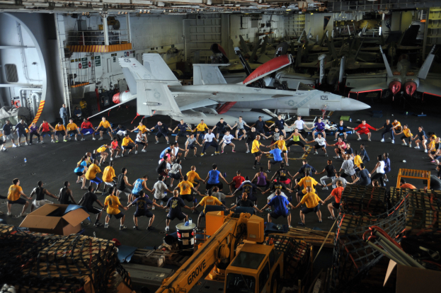 Eine Gruppe von Menschen auf einem Flugzeugträger umgeben von Kisten, Maschinen und anderen Gegenständen, mit Flugzeugen und anderen Fluggeräten im Hintergrund.