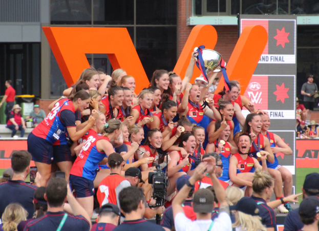 Eine Gruppe von Frauenfußballerinnen in blauen und roten Uniformen feiert mit einem Pokal vor einer jubelnden Menge auf einem Rasenfeld, mit Zuschauern und beschrifteten Tafeln im Hintergrund.