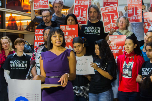 Eine Black-Lives-Matter-Aktivistin spricht an einem Podium mit Mikrofon zu einer Menge, die Schilder hält, mit Lichtern und Gegenständen im Hintergrund.