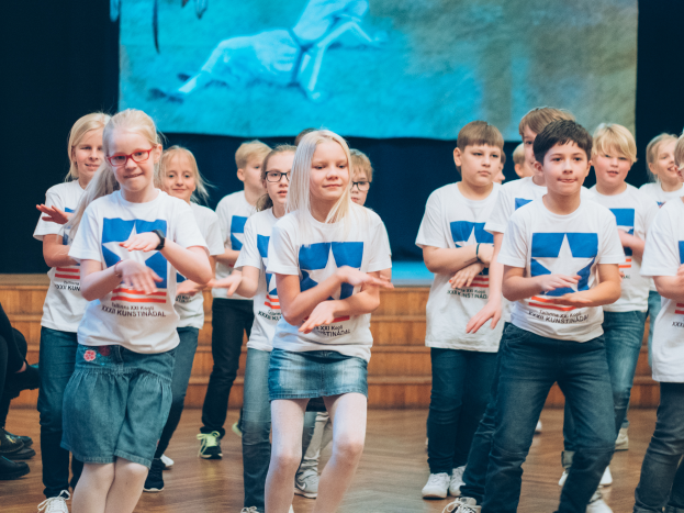 Eine Gruppe von Kindern in weißen Hemden und blauen Shorts, einige mit Brille, tanzt auf einer Bühne mit Zuschauern auf der linken Seite und einem Bildschirm im Hintergrund, der an einem Wettbewerb teilnimmt.