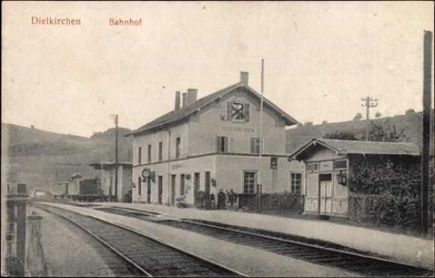 Schwarz-weiß-Foto eines Bahnhofs in Dielkirchen, Deutschland, mit einem Zug auf den Gleisen, umgeben von Häusern, Pfählen, Bäumen, Pflanzen und Menschen, Bergen im Hintergrund und einem klaren Himmel.