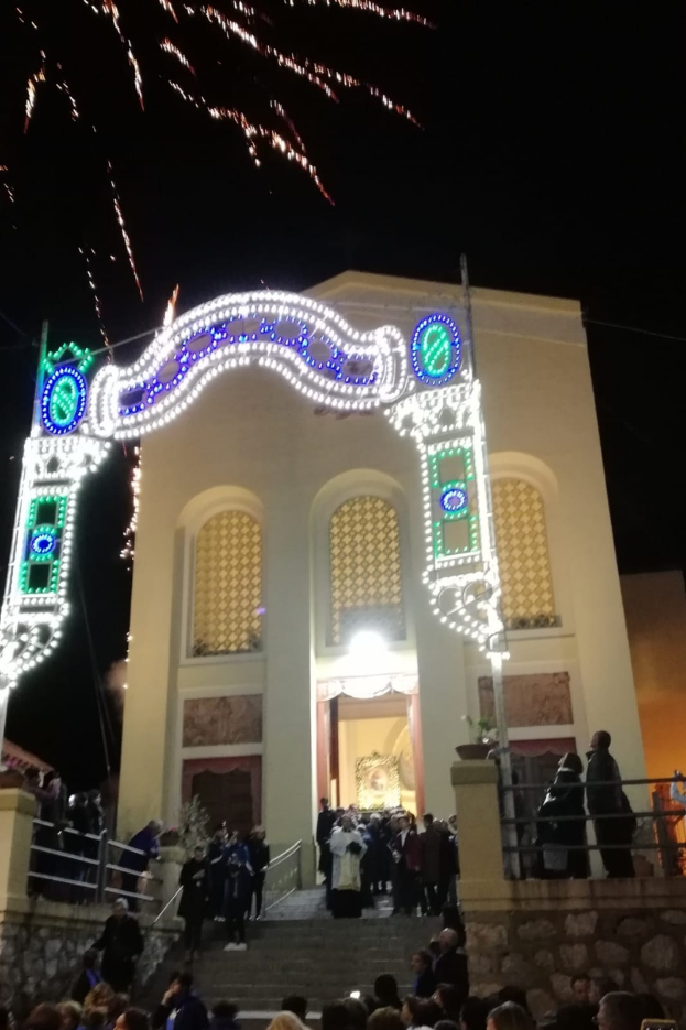 Menschen, die vor einem Gebäude mit einem bogenförmigen, mit Lichtern geschmückten Eingang, Treppen mit Geländern und Topfpflanzen stehen und Silvester mit Feuerwerk im Hintergrund feiern.