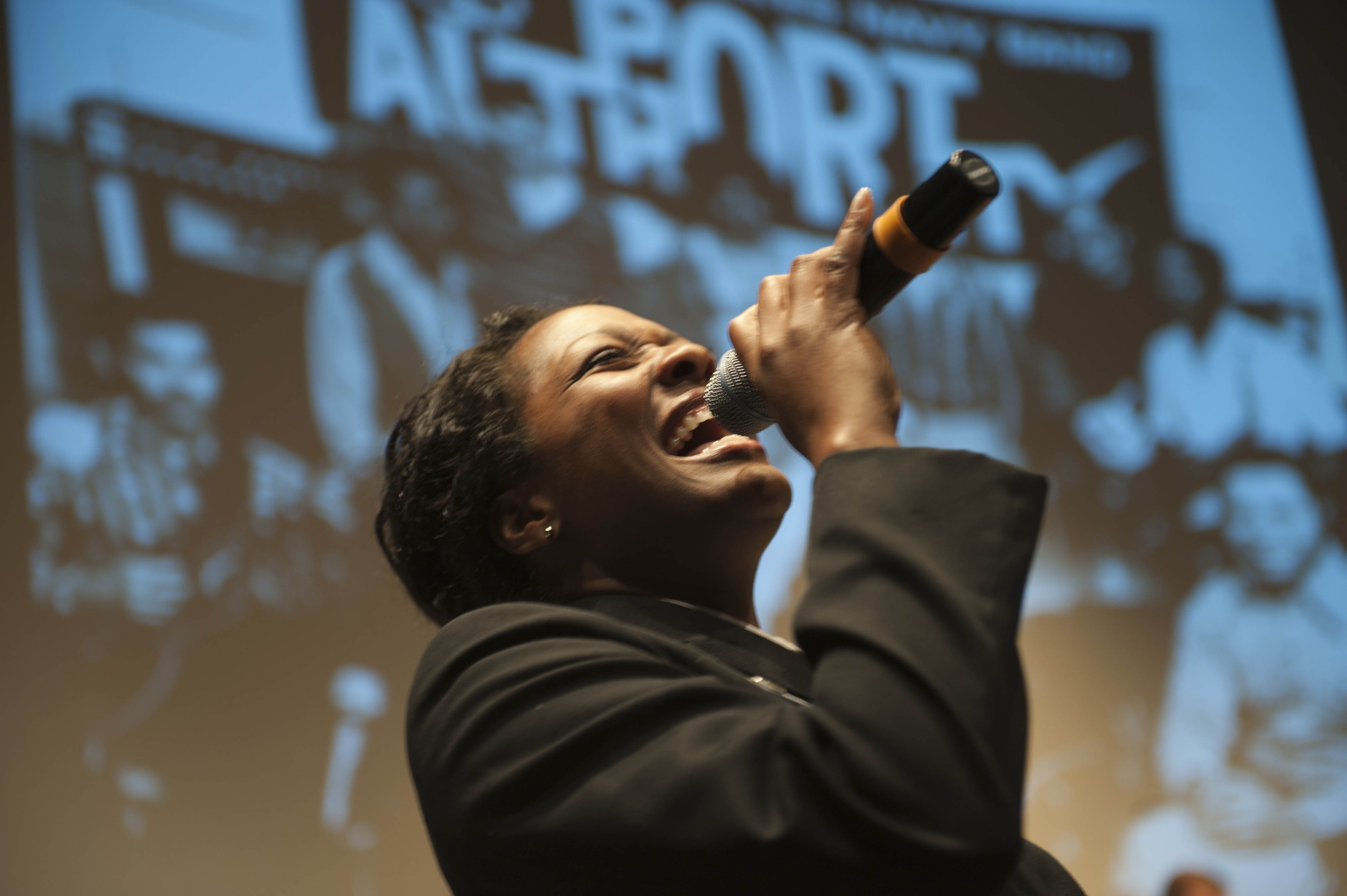 Eine Frau in einem schwarzen Kleid mit einem Dutt singt leidenschaftlich in ein Mikrofon auf der Bühne, ihr Gesicht von Scheinwerfern beleuchtet, die Augen vor Emotion geschlossen.