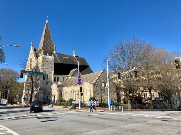 Große Kirche mit Turm an der Ecke einer Straße, beschriftet als St. Lukes Episkopalkirche, umgeben von Gebäuden, Straßenmöblierung, Fahrzeugen, Passanten, Grünflächen und einem klaren Himmel.