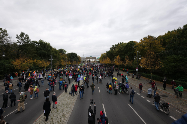 Eine große Gruppe von Menschen marschiert eine baumbestandene Straße mit Laternenpfählen entlang, hält Kameras, mit einem Gebäude und einem klaren Himmel im Hintergrund, was auf eine Demonstration in Berlin hindeutet.