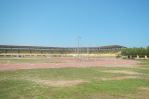 Ein großes Stadion mit einer zentralen Laufbahn, umgeben von grünem Gras und Bäumen auf der rechten Seite, mit Pfosten und einem klaren blauen Himmel im Hintergrund.