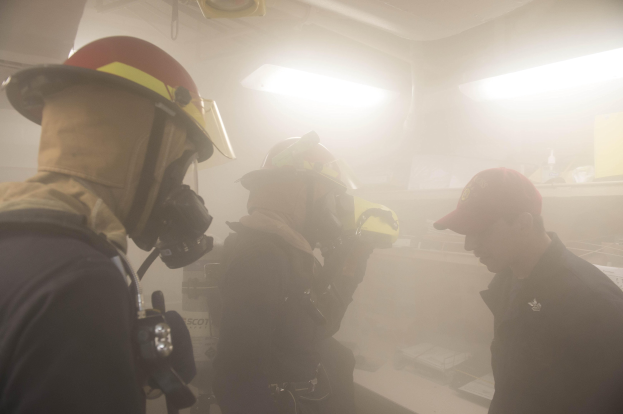 Feuerwehrleute in Helmen mit Rauch aus dem Mund, einer hält eine Kamera, während einer Trainingsrunde mit Lichtern und Gegenständen im Hintergrund.