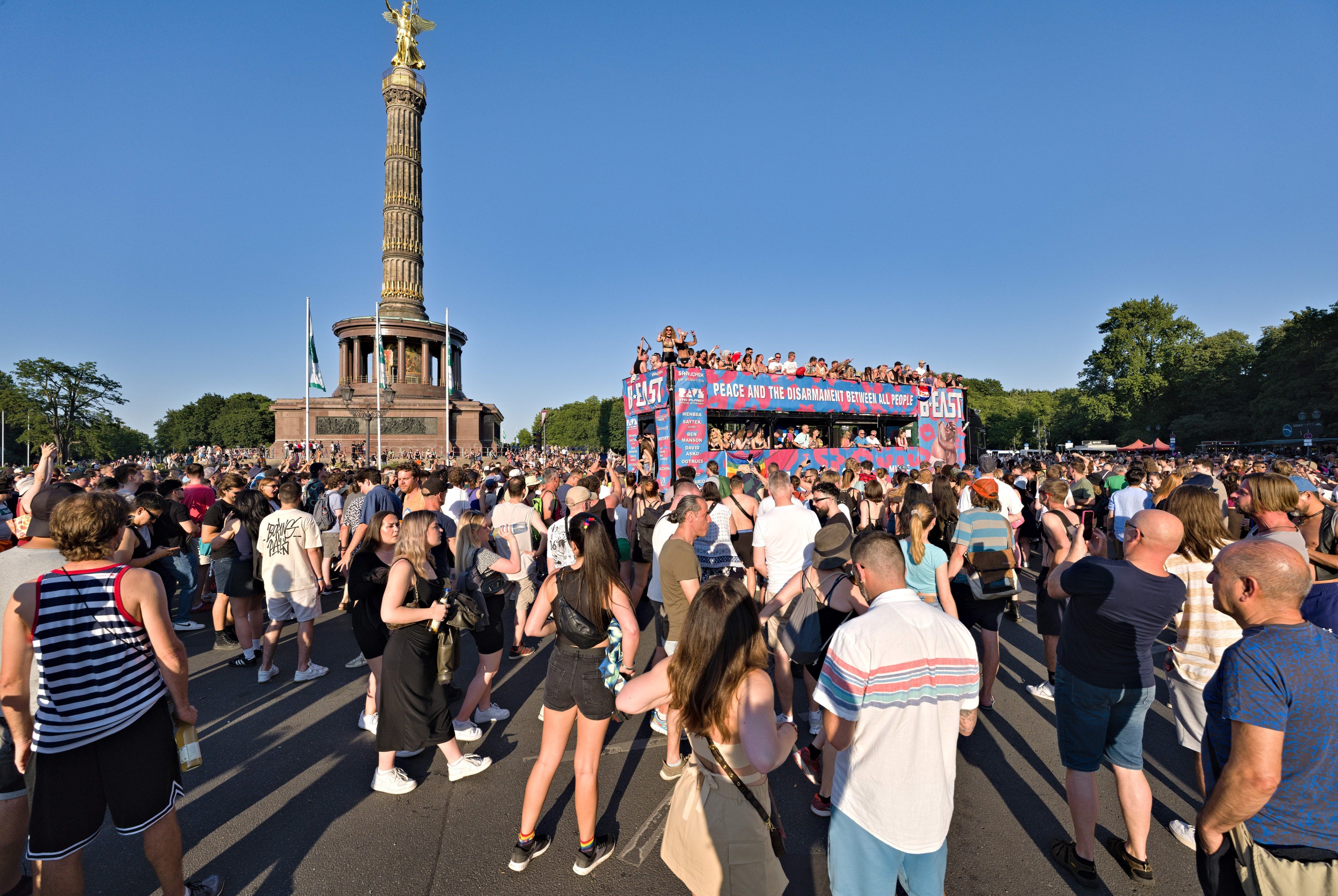 Große Menschenmenge vor einem Berliner Denkmal mit Fahnen, einem Gebäude mit Säulen, einer Statue und Inschriften, vor einem Baum und einem klaren blauen Himmel.