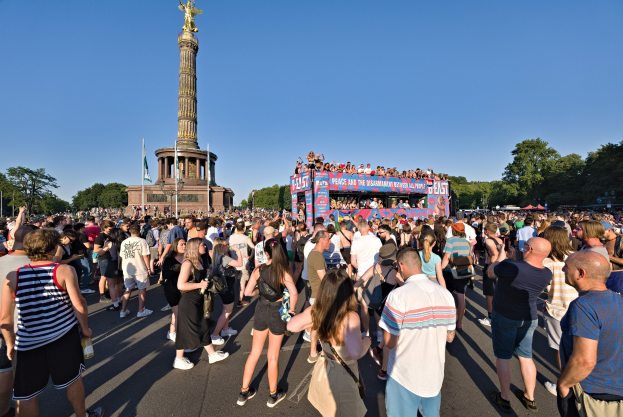 Große Menschenmenge vor einem Berliner Denkmal mit Fahnen, einem Gebäude mit Säulen, einer Statue und Inschriften, vor einem Baum und einem klaren blauen Himmel.