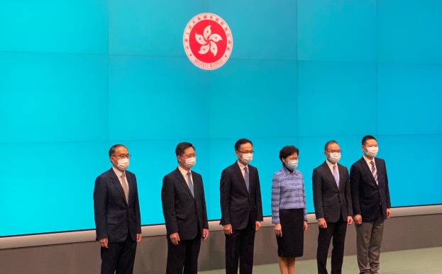 Eine Gruppe maskierter Individuals steht nebeneinander vor einer blauen Wand mit einem Logo und nimmt an einer Pressekonferenz in Hongkong teil.