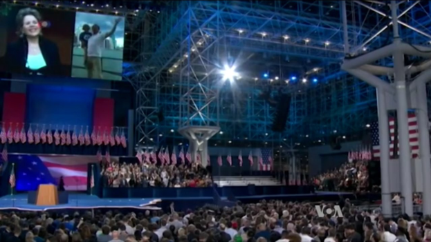 Eine große Menschenmenge sitzt vor einer Bühne mit einem Podium, Mikrofon, Fahnen, einem Bildschirm und Beleuchtungsausrüstung und wartet auf eine Präsidentenrede.