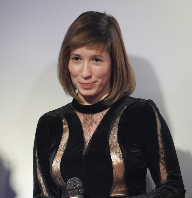 Eine Frau in einem schwarzen Kleid hält ein Mikrofon und steht vor einer Wand, wobei sie selbstbewusst und entschlossen wirkt.