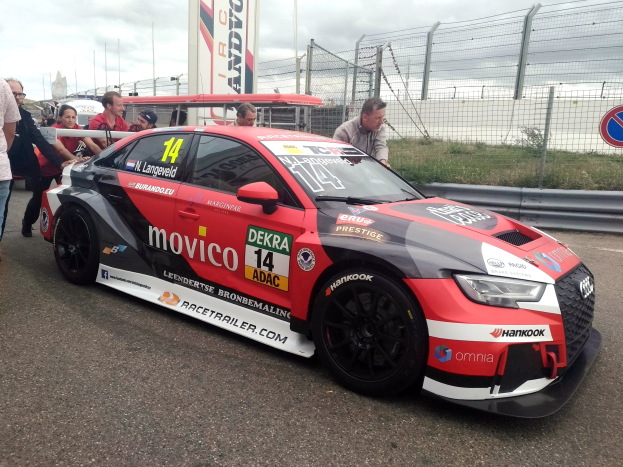 Audi RS3 DTM-Rennwagen auf einer Straße mit einer Gruppe von Menschen, einem Zaun, Gras, einem Schild, einer Fahne mit Text, Pfosten und einem bewölkten Himmel im Hintergrund.