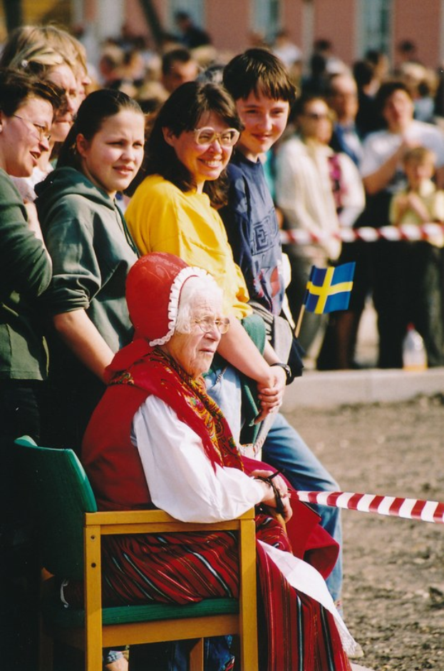 Alte Frau in einem Stuhl sitzend, die eine Menge ansieht, mit einer Schleife im Vordergrund und einem Gebäude im Hintergrund