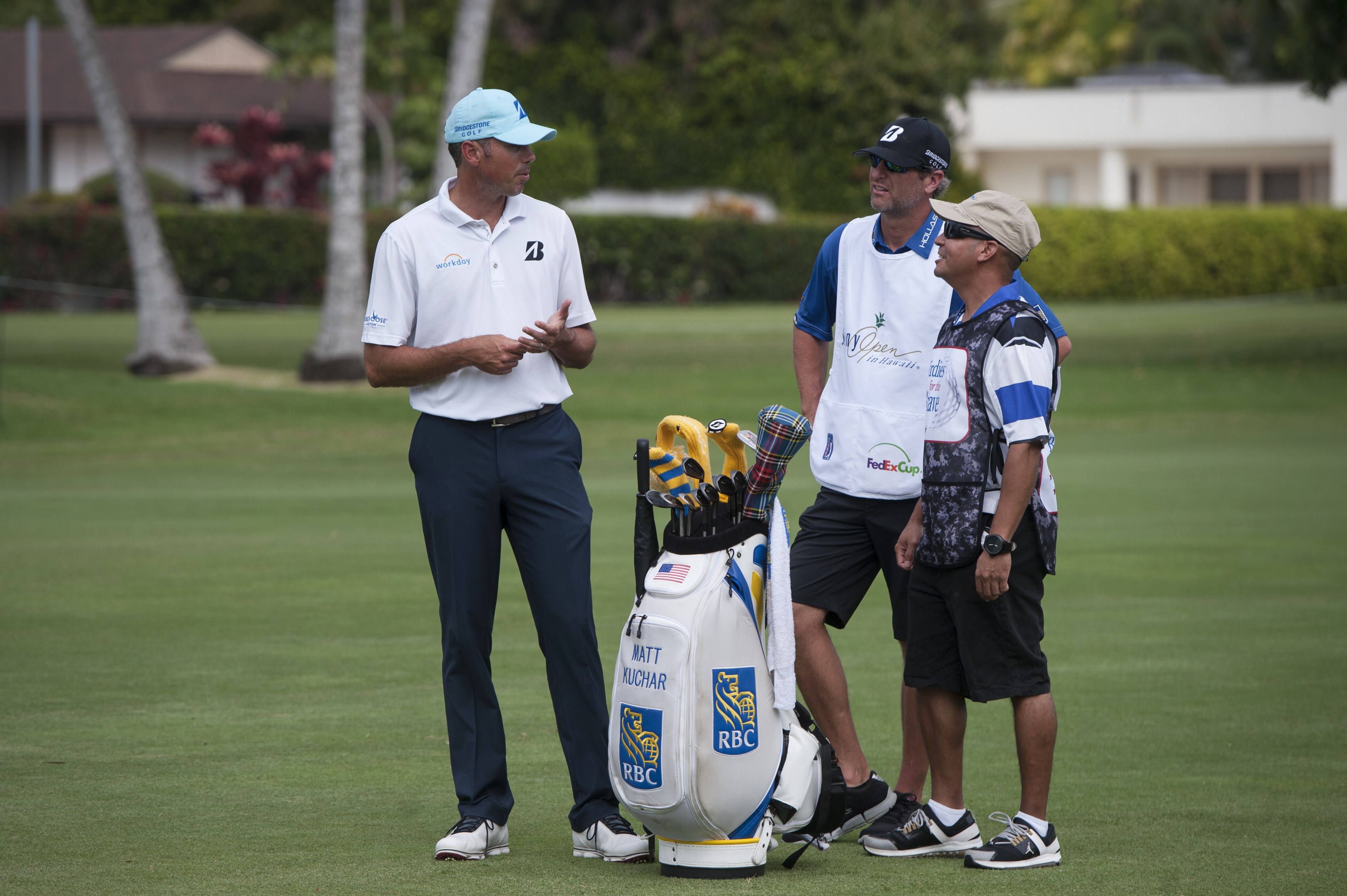Mannergruppe auf einem grünen Feld mit Golfbag und -schlägern im Vordergrund, Bäume und Gebäude im Hintergrund.