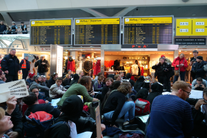 Eine große Gruppe von Menschen versammelt sich in einem Flughafen, einige sitzen mit Taschen und Papieren, andere stehen, mit Texttafeln, Schaufensterpuppen in Kleidern und Deckenleuchten im Hintergrund, was auf eine Protestaktion hindeutet.