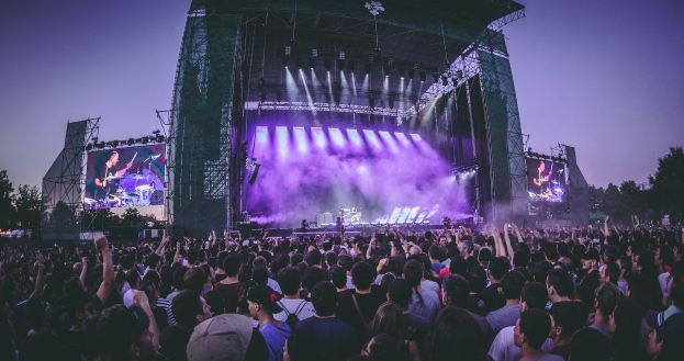 Eine Menschenmenge bei einem Musikfestival mit einer Bühne, auf der Musiker Instrumente spielen, beleuchtet von Scheinwerfern und umgeben von Bildschirmen, vor einer Kulisse aus einem Gebäude, Bäumen und einem klaren blauen Himmel.