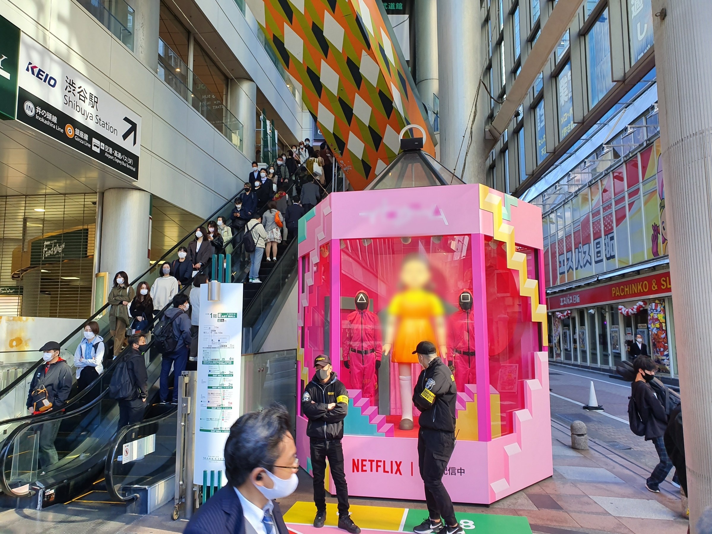 Eine Gruppe von Menschen steht vor einem Gebäude mit Glaswänden, Säulen und Texttafeln, einige tragen Masken, während eine Rolltreppe mit Menschen daneben verläuft; ein pinker Kasten in der Mitte zeigt Schaufensterpuppen und trägt "Netflix's new Netflix booth in Tokyo."