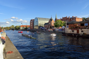 Eine Gruppe von Menschen rudert in einem Boot einen Fluss in Kopenhagen, Dänemark, hinunter, während Zuschauer auf einer Mauer links und Gebäude, Bäume und ein bewölkter Himmel im Hintergrund zu sehen sind.