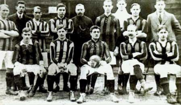 Schwarzes und weißes Foto einer Fussballmannschaft, einige stehen und einige sitzen, vor einer Wand mit dem Text "Ballymena Football Club" unten.