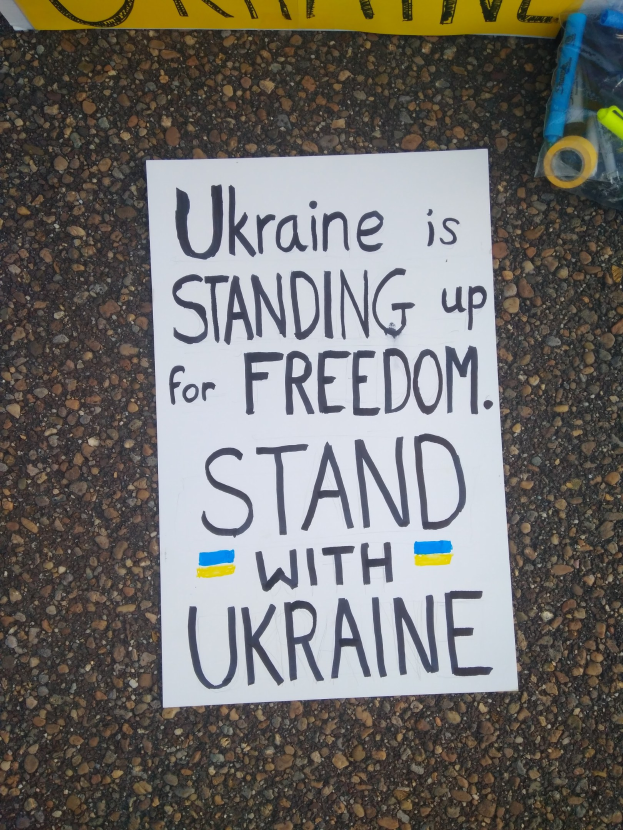 Weißer Zettel mit dem Text "Ukraine steht für Freiheit auf, stellt dich mit Ukraine" auf dem Boden vor einer gelben Fahne, mit einer Plastikhülle mit verschiedenen Gegenständen rechts daneben.