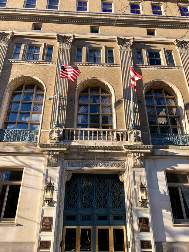 Außenansicht des Plaza Hotels in New York City, mit Glasfenstern, Türen, Säulen, Geländern, Leuchten, Texttafeln und Flaggen vor einem Himmelhintergrund.