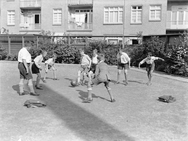 Schwarzes und weißes Foto von jungen Jungs, die auf einem Rasenfeld Fußball spielen, mit einem Ball in der Mitte, Gebäuden mit Fenstern, Geländern und Balkonen im Hintergrund und Pflanzen und Bäumen, die die Gegend umgeben.
