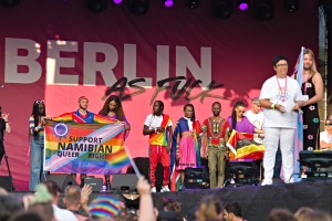 Gruppe von Menschen auf einer Bühne mit einem Banner, auf dem "Berlin as Fuck" steht, mit Lautsprechern, Mikrofonstativen und einer feiernden Menge unten