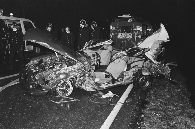 Schwarz-weiß-Foto eines auf der Straße liegenden Wagens mit mehreren Personen, einige mit Helmen, drumherum und einem Fahrzeug im Hintergrund.