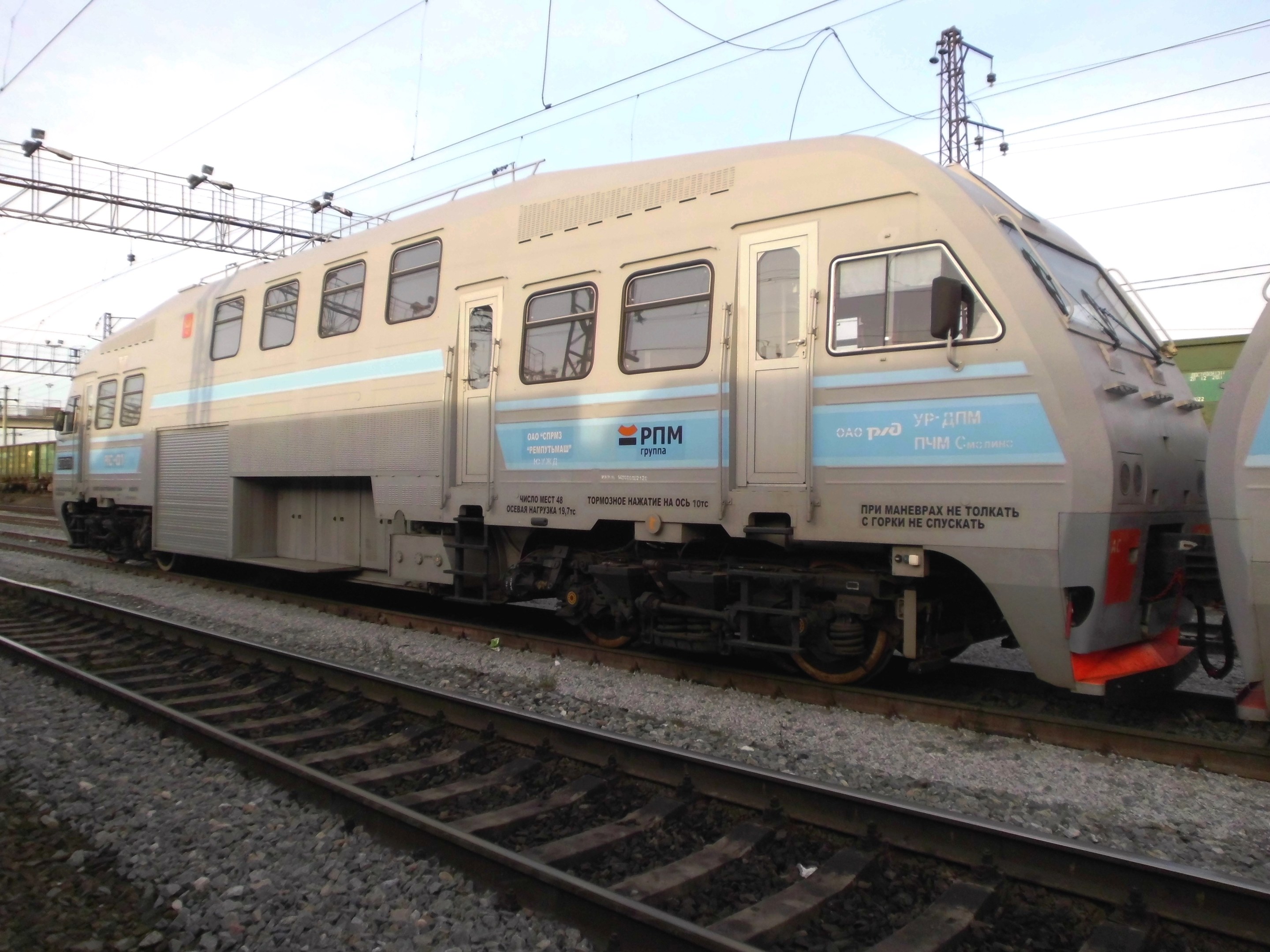 Diesellokomotive auf Schienen neben einem anderen Zug fahrend, mit Strommasten und Kabeln im Hintergrund unter einem klaren blauen Himmel.