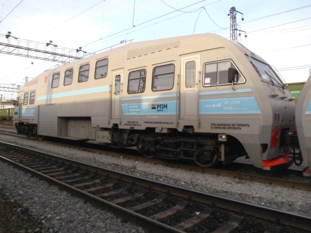 Diesellokomotive auf Schienen neben einem anderen Zug fahrend, mit Strommasten und Kabeln im Hintergrund unter einem klaren blauen Himmel.