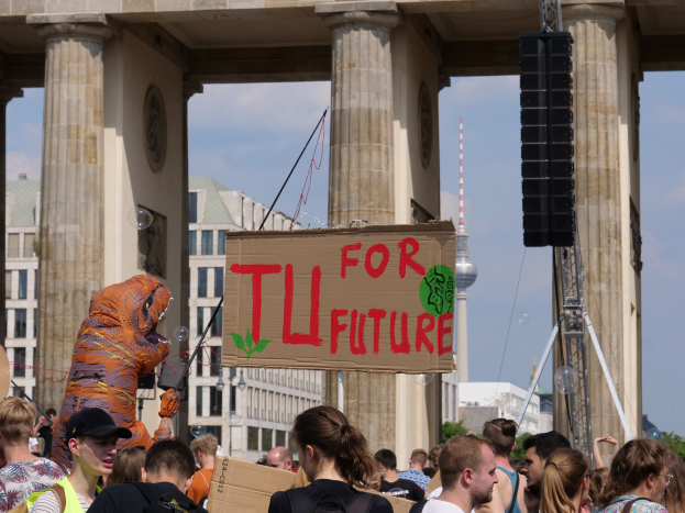Eine Gruppe von Menschen steht vor dem Brandenburger Tor in Berlin, Deutschland, wobei eine Person in Kostüm mit einem Schild und andere mit Taschen zu sehen sind, vor einem Hintergrund von Gebäuden, Bäumen und einem Turm unter einem bewölkten Himmel.