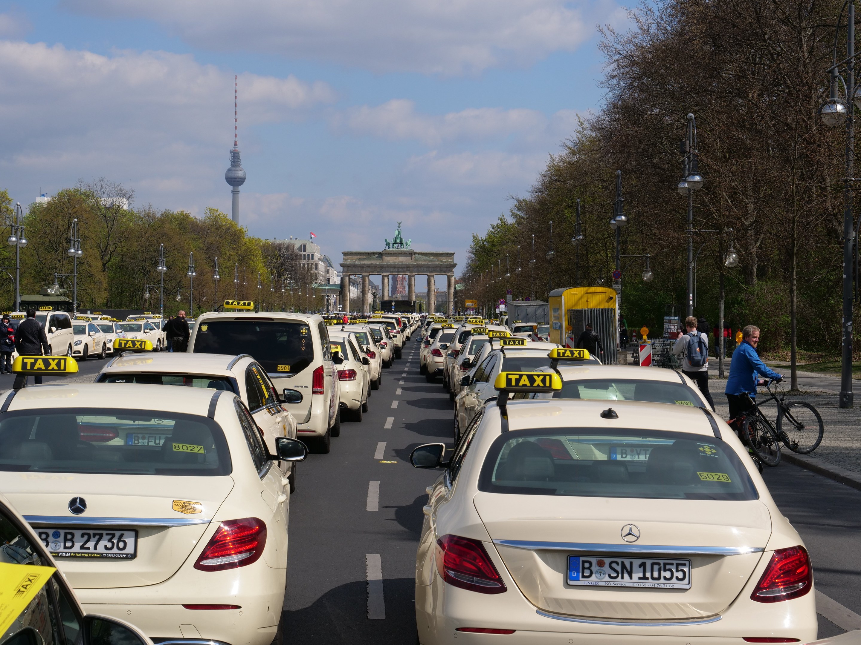 Eine lange Reihe von Taxis, die an einer belebten Straße in Berlin, Deutschland, geparkt sind, mit Fahrradfahrern und Füßgängern auf dem Gehweg, flankiert von Bäumen und Laternenmästen und Gebäuden, einem Bogen und einem Turm im Hintergrund bei einem bewölktem Himmel.