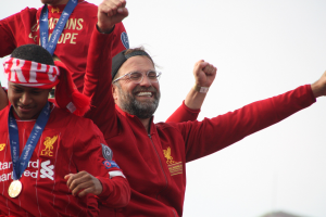 Liverpool-Trainer Jürgen Klopp feiert mit Spielern und Staff nach dem Gewinn der UEFA Champions League, trägt eine rote Jacke und hat zwei Medaillengewinner neben sich vor einem Himmel-Hintergrund.