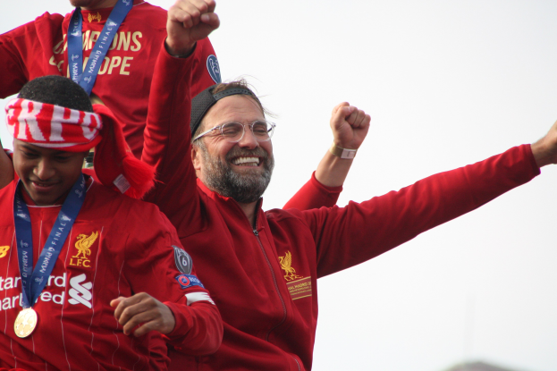 Liverpool-Trainer Jürgen Klopp feiert mit Spielern und Staff nach dem Gewinn der UEFA Champions League, trägt eine rote Jacke und hat zwei Medaillengewinner neben sich vor einem Himmel-Hintergrund.