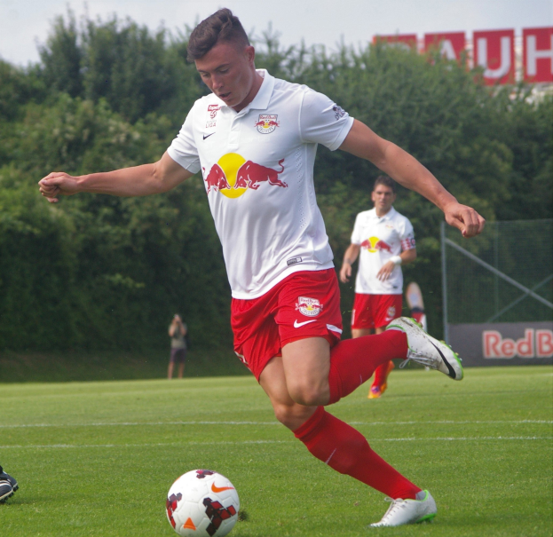 Ein professioneller Fußballspieler in einem roten Trikot kickt einen Fußball auf einem grünen Feld mit Bäumen und einem klaren blauen Himmel im Hintergrund, mit einer Tafel mit der Aufschrift "RB Leipzig" im Bild.