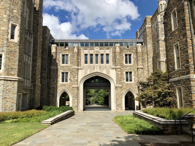 Großes steinernes Universitätsgebäude mit gewölbtem Eingang, umgeben von grünem Gras, Pflanzen, Bäumen und einem teilweise bewölkten Himmel.