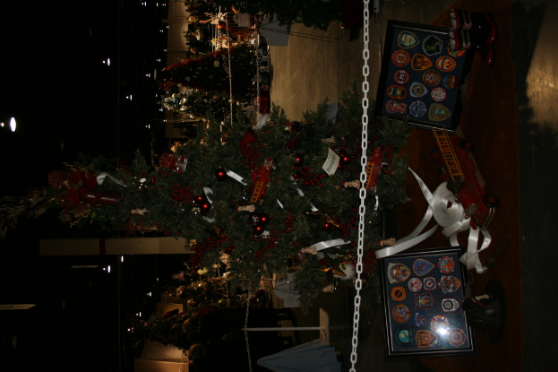 Ein geschm├╝ckter Weihnachtsbaum mit Geschenken, Rahmen und Spielzeugen auf einer Plattform, mit Menschen, weiteren Weihnachtsb├Ąumen und Lichtern im Hintergrund.