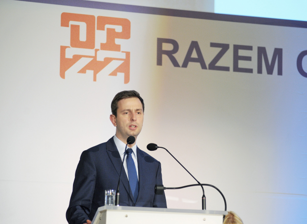 Ein Mann im Anzug steht an einem Podium mit einem Glas Wasser und zwei Mikrofonen und spricht als CEO von Razem Global, mit einem Banner mit der Aufschrift "Razem Global" im Hintergrund.