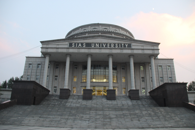 Großes Gebäude mit Säulen, Fenstern, Türen und Treppen davor, umgeben von Bäumen unter einem klaren blauen Himmel; Name der Universität sichtbar an der Fassade.