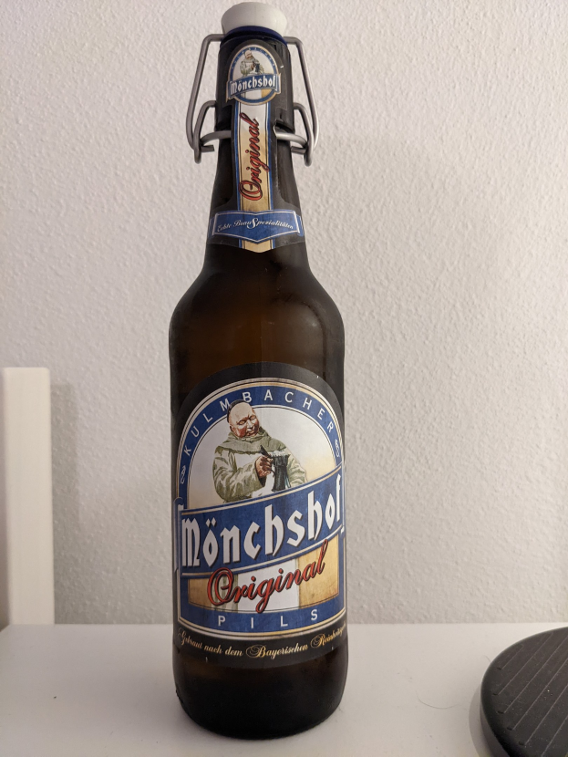 Eine Flasche Münchner Bier mit einem Aufkleber darauf auf einem weißen Tisch neben einem schwarzen Objekt, mit einer weißen Wand im Hintergrund.