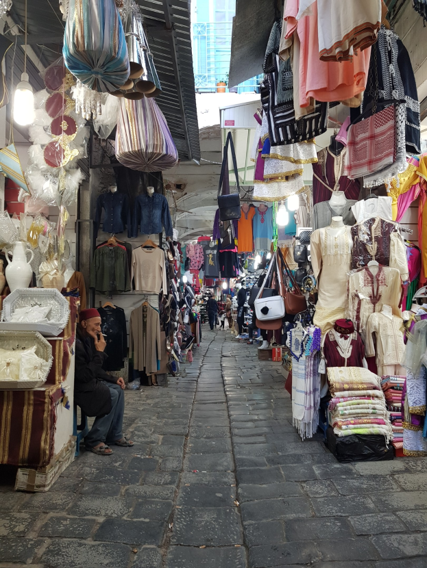 Ein belebter Straßenmarkt in der Altstadt von Jerusalem mit Ständen, die Kleidung, Taschen, Lampen und andere Artikel verkaufen, während Menschen durch die Gassen schlendern und das Angebot inspizieren, mit einem Gebäude im Hintergrund.