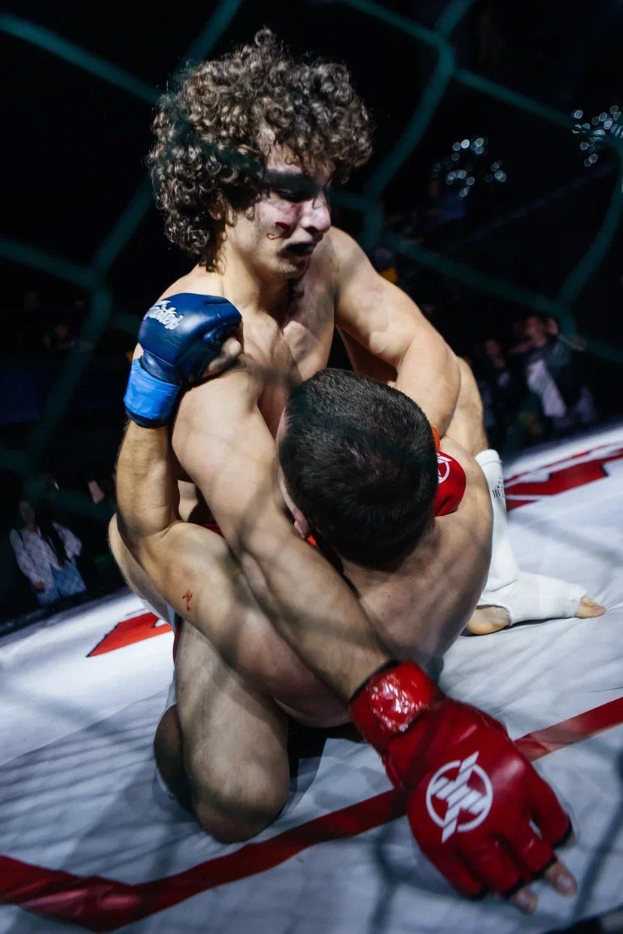 Zwei Männer in einem MMA-Ring im Kampf, einer setzt einen Tritt gegen den anderen, umgeben von einem Netz mit dunklem Hintergrund.