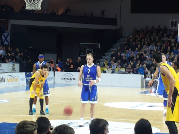 Eine Gruppe von Männern in blauen und gelben Uniformen spielt Basketball auf einem Court mit einem Netz und einer Wand im Hintergrund, während Zuschauer von den Rängen zuschauen während eines 7days Eurocup-Spiels zwischen Fenerbahce und Real Madrid.